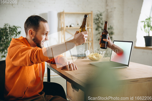 Image of Young man drinking beer during meeting friends on virtual video call. Distance online meeting, chat together on laptop at home.