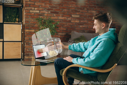 Image of Young man drinking beer during meeting friends on virtual video call. Distance online meeting, chat together on laptop at home.