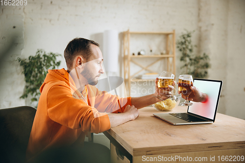 Image of Young man drinking beer during meeting friends on virtual video call. Distance online meeting, chat together on laptop at home.