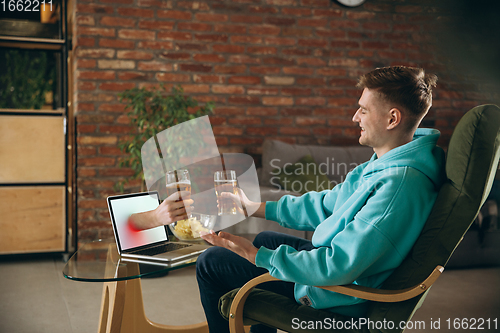 Image of Young man drinking beer during meeting friends on virtual video call. Distance online meeting, chat together on laptop at home.