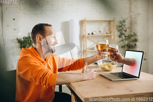 Image of Young man drinking beer during meeting friends on virtual video call. Distance online meeting, chat together on laptop at home.