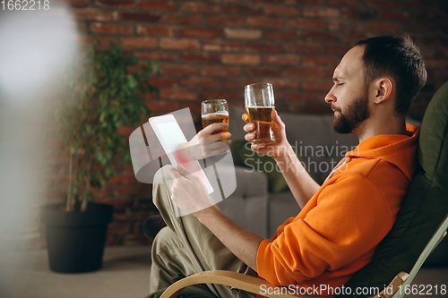 Image of Young man drinking beer during meeting friends on virtual video call. Distance online meeting, chat together on tablet at home.