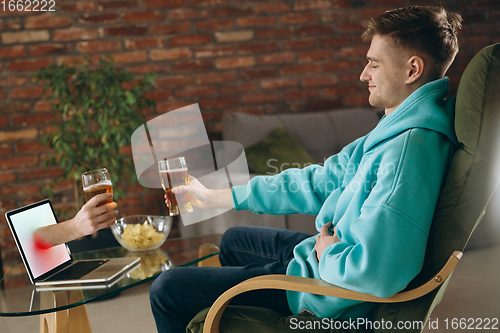 Image of Young man drinking beer during meeting friends on virtual video call. Distance online meeting, chat together on laptop at home.