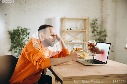 Image of Young man drinking beer during meeting friends on virtual video call. Distance online meeting, chat together on laptop at home.