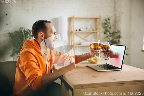 Image of Young man drinking beer during meeting friends on virtual video call. Distance online meeting, chat together on laptop at home.