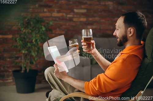 Image of Young man drinking beer during meeting friends on virtual video call. Distance online meeting, chat together on tablet at home.