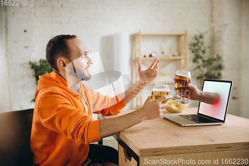 Image of Young man drinking beer during meeting friends on virtual video call. Distance online meeting, chat together on laptop at home.