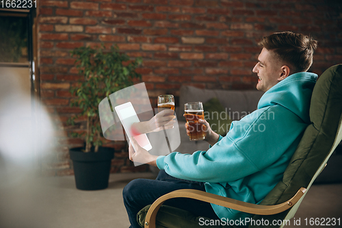 Image of Young man drinking beer during meeting friends on virtual video call. Distance online meeting, chat together on tablet at home.
