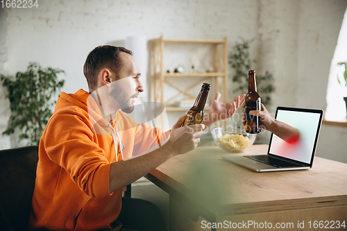 Image of Young man drinking beer during meeting friends on virtual video call. Distance online meeting, chat together on laptop at home.