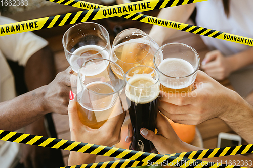 Image of Close up hands clinking glasses of beer at bar with bounding tapes Lockdown, Coronavirus, Quarantine, Warning - closing bars and nightclubs during pandemic