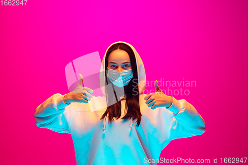 Image of Caucasian beautiful woman\'s portrait isolated on pink studio background in multicolored neon light. Wearing face mask.