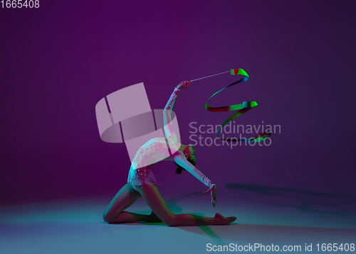 Image of Young female athlete, rhythmic gymnastics artist on purple background with neon light. Beautiful girl practicing with equipment. Grace in performance.