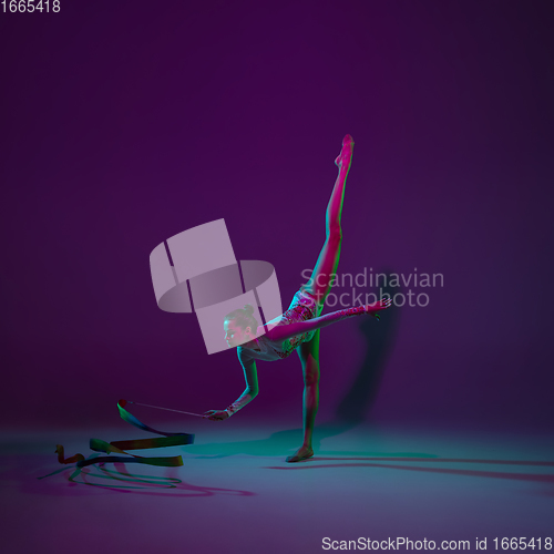 Image of Young female athlete, rhythmic gymnastics artist on purple background with neon light. Beautiful girl practicing with equipment. Grace in performance.