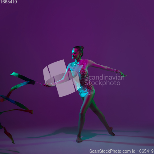 Image of Young female athlete, rhythmic gymnastics artist on purple background with neon light. Beautiful girl practicing with equipment. Grace in performance.
