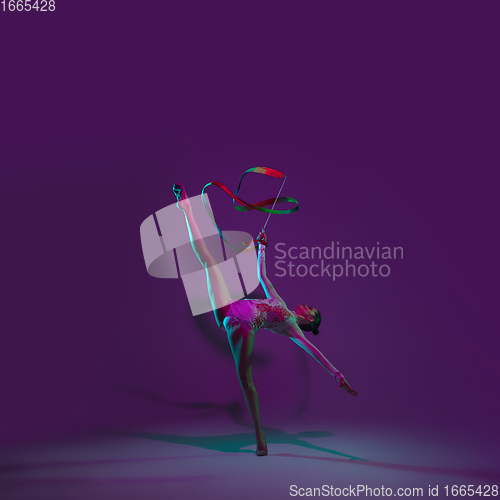 Image of Young female athlete, rhythmic gymnastics artist on purple background with neon light. Beautiful girl practicing with equipment. Grace in performance.