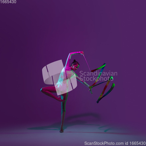 Image of Young female athlete, rhythmic gymnastics artist on purple background with neon light. Beautiful girl practicing with equipment. Grace in performance.