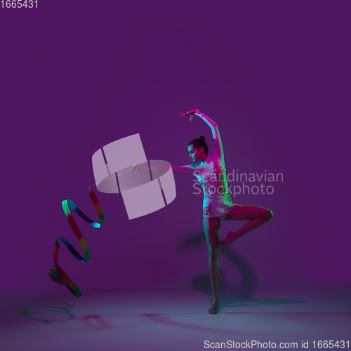 Image of Young female athlete, rhythmic gymnastics artist on purple background with neon light. Beautiful girl practicing with equipment. Grace in performance.