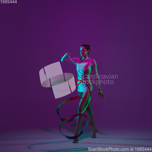 Image of Young female athlete, rhythmic gymnastics artist on purple background with neon light. Beautiful girl practicing with equipment. Grace in performance.