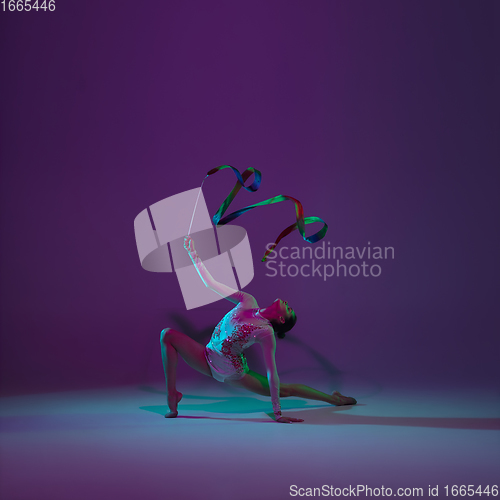 Image of Young female athlete, rhythmic gymnastics artist on purple background with neon light. Beautiful girl practicing with equipment. Grace in performance.