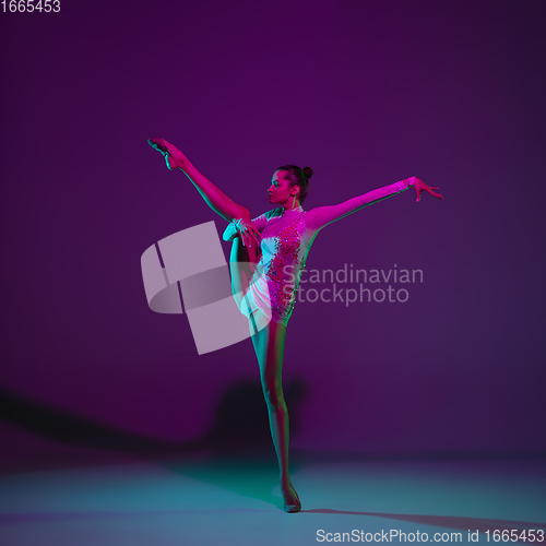 Image of Young female athlete, rhythmic gymnastics artist on purple background with neon light. Beautiful girl practicing with equipment. Grace in performance.