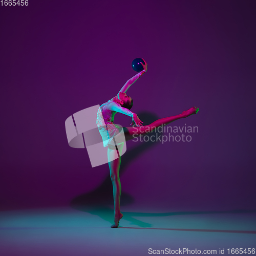 Image of Young female athlete, rhythmic gymnastics artist on purple background with neon light. Beautiful girl practicing with equipment. Grace in performance.