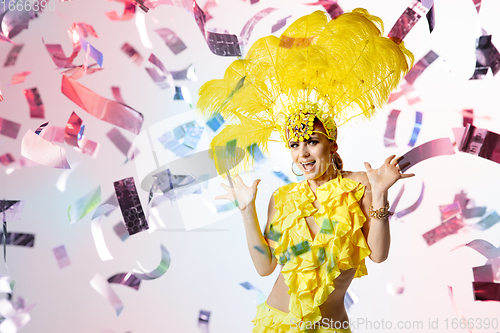 Image of Beautiful young woman in carnival, stylish masquerade costume with feathers dancing on white studio background with shining confetti