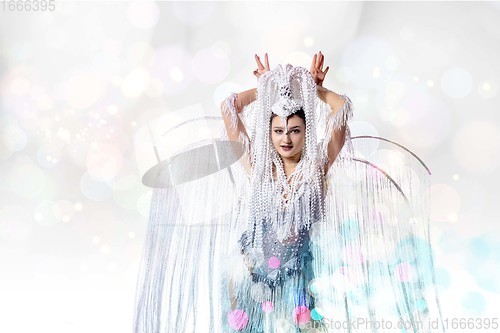 Image of Beautiful young woman in carnival, stylish masquerade costume with feathers dancing on white studio background with shining confetti