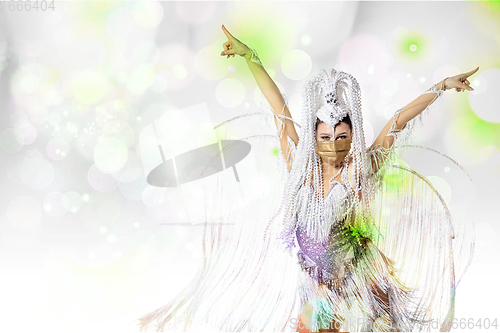 Image of Beautiful young woman in carnival, stylish masquerade costume with feathers dancing on white studio background with shining bokeh