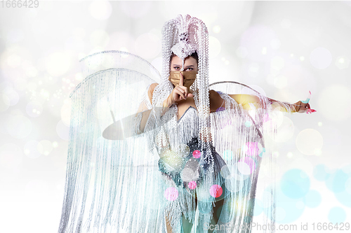 Image of Beautiful young woman in carnival, stylish masquerade costume with feathers dancing on white studio background with shining bokeh