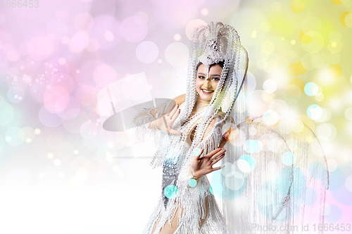 Image of Beautiful young woman in carnival, stylish masquerade costume with feathers dancing on white studio background with shining confetti