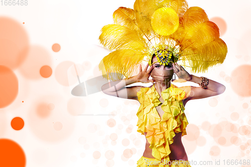 Image of Beautiful young woman in carnival, stylish masquerade costume with feathers dancing on white studio background with shining bokeh