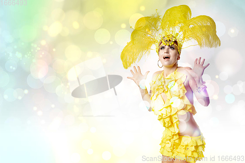 Image of Beautiful young woman in carnival, stylish masquerade costume with feathers dancing on white studio background with shining bokeh