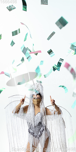 Image of Beautiful young woman in carnival, stylish masquerade costume with feathers dancing on white studio background with shining confetti