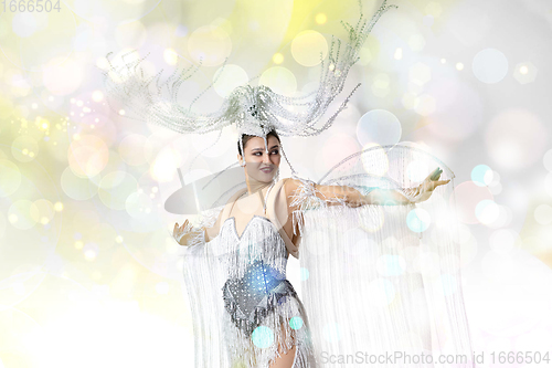 Image of Beautiful young woman in carnival, stylish masquerade costume with feathers dancing on white studio background with shining confetti