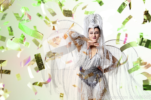 Image of Beautiful young woman in carnival, stylish masquerade costume with feathers dancing on white studio background with shining confetti