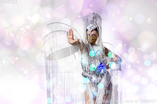 Image of Beautiful young woman in carnival, stylish masquerade costume with feathers dancing on white studio background with shining bokeh