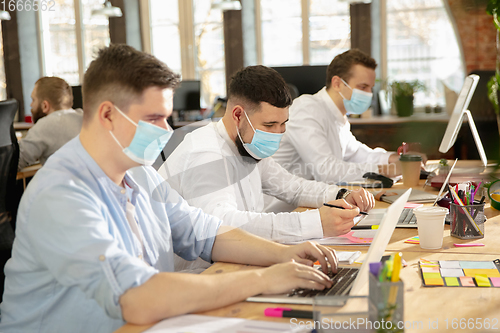 Image of Young caucasian colleagues working together in a office using modern devices and gadgets during quarantine. Wearing protective face masks