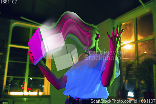 Image of Cinematic portrait of stylish woman in neon lighted interior using a tablet. The face is smeared, sucked into the screen. The concept of social network dependency