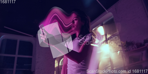 Image of Cinematic portrait of stylish woman in neon lighted interior using a smartphone. The face is smeared, sucked into the phone. The concept of social network dependency