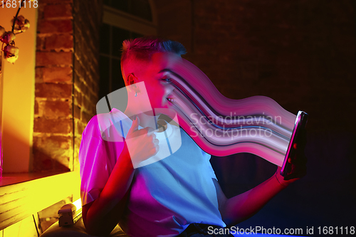 Image of Cinematic portrait of stylish woman in neon lighted interior using a smartphone. The face is smeared, sucked into the screen. The concept of social network dependency