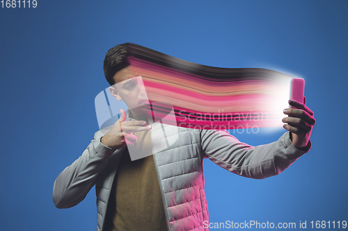 Image of Cinematic portrait of stylish man in neon lighted interior using a smartphone. The face is smeared, sucked into the phone. The concept of social network dependency