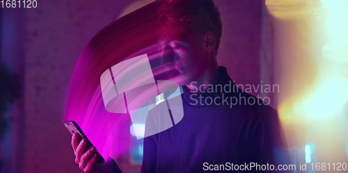 Image of Cinematic portrait of stylish man in neon lighted interior using a smartphone. The face is smeared, sucked into the phone. The concept of social network dependency