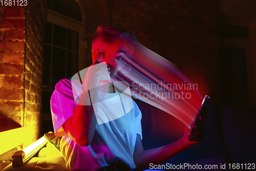 Image of Cinematic portrait of stylish woman in neon lighted interior using a smartphone. The face is smeared, sucked into the phone. The concept of social network dependency