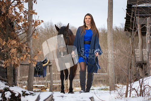 Image of A girl in a blue dress walks with a horse through the winter forest and old wooden ruins