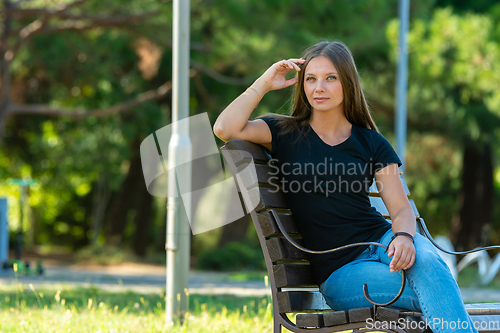 Image of A beautiful girl in casual clothes sits on a bench in a beautiful green park, the girl looks into the frame