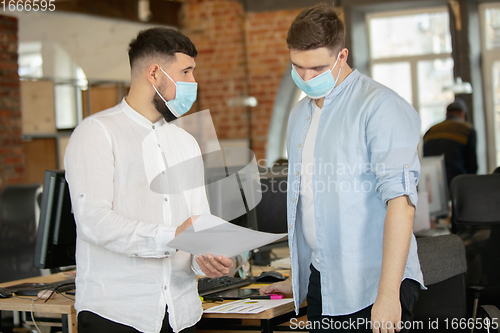 Image of Young caucasian colleagues working together in a office using modern devices and gadgets during quarantine. Wearing protective face masks