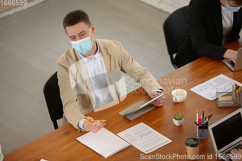 Image of High angle view colleagues working together in face masks during quarantine in a office using modern devices and gadgets during creative meeting