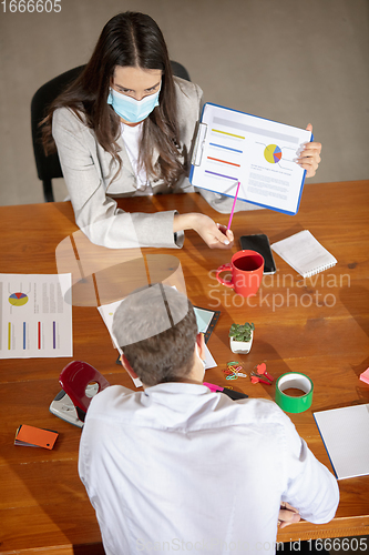 Image of High angle view colleagues working together in face masks during quarantine in a office using modern devices and gadgets during creative meeting