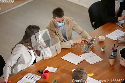 Image of High angle view colleagues working together in face masks during quarantine in a office using modern devices and gadgets during creative meeting