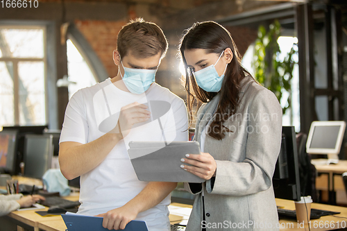 Image of Young caucasian colleagues working together in a office using modern devices and gadgets during quarantine. Wearing protective face masks
